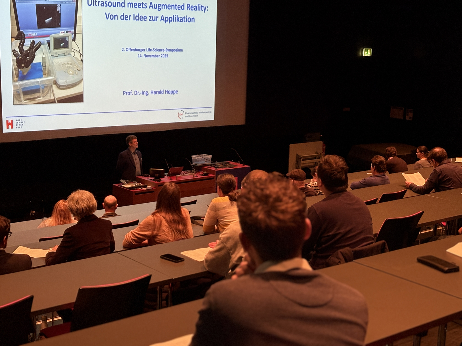 Ein Mann steht in einem Hörsaal vor einer Leinwand an einem Rednerpult.