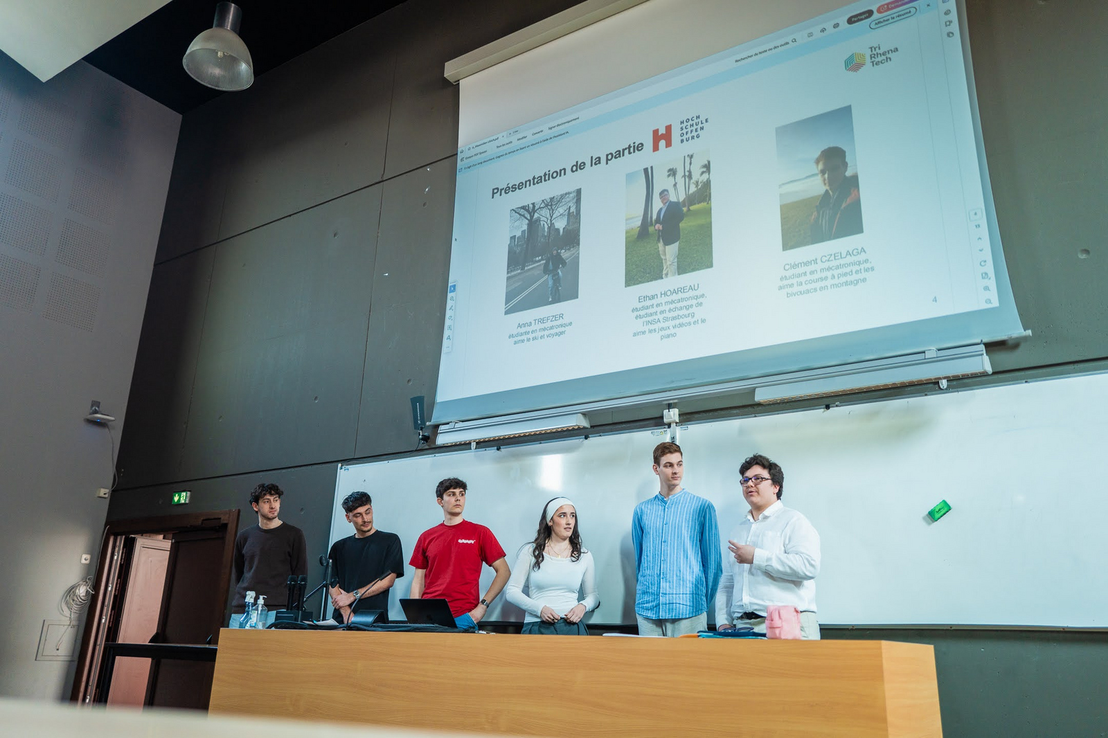Sechs junge Leute stehen an einem langen Rednerpult vor einer Leinwand und stellen sich vor