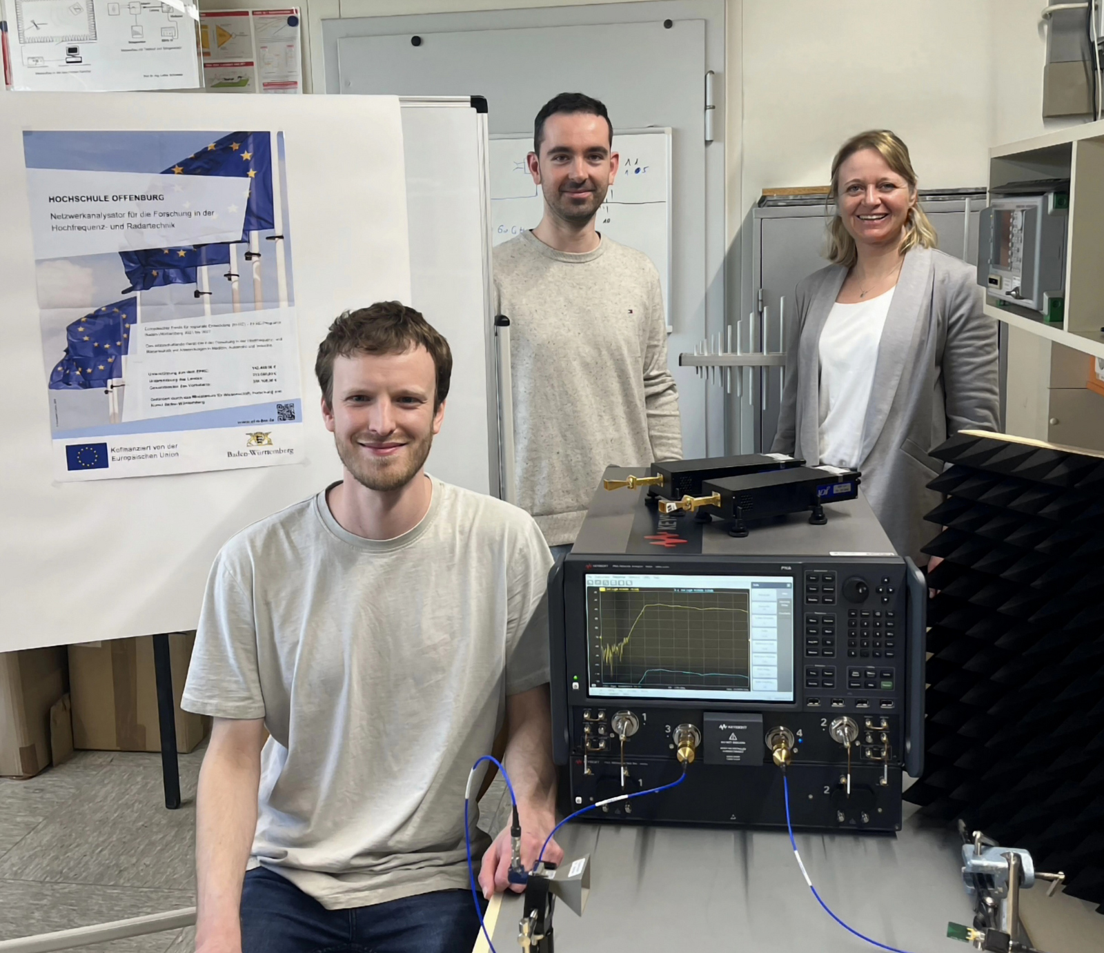 zwei Männer und eine Frau sitzen beziehunsgweise stehen um den Netzwerkanalysator, der auf einem Tisch steht, herum