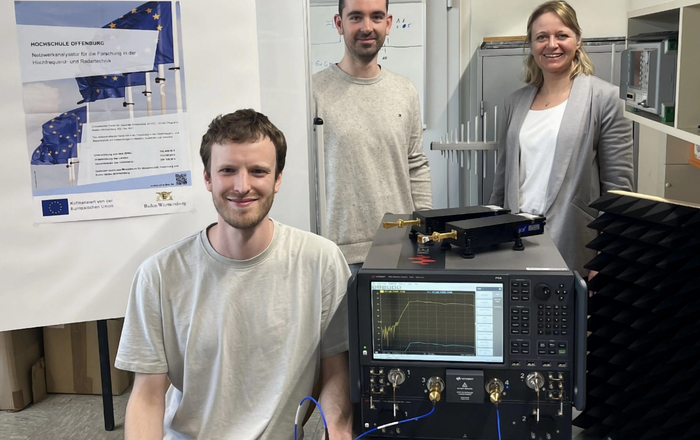 zwei Männer und eine Frau sitzen beziehunsgweise stehen um den Netzwerkanalysator, der auf einem Tisch steht, herum