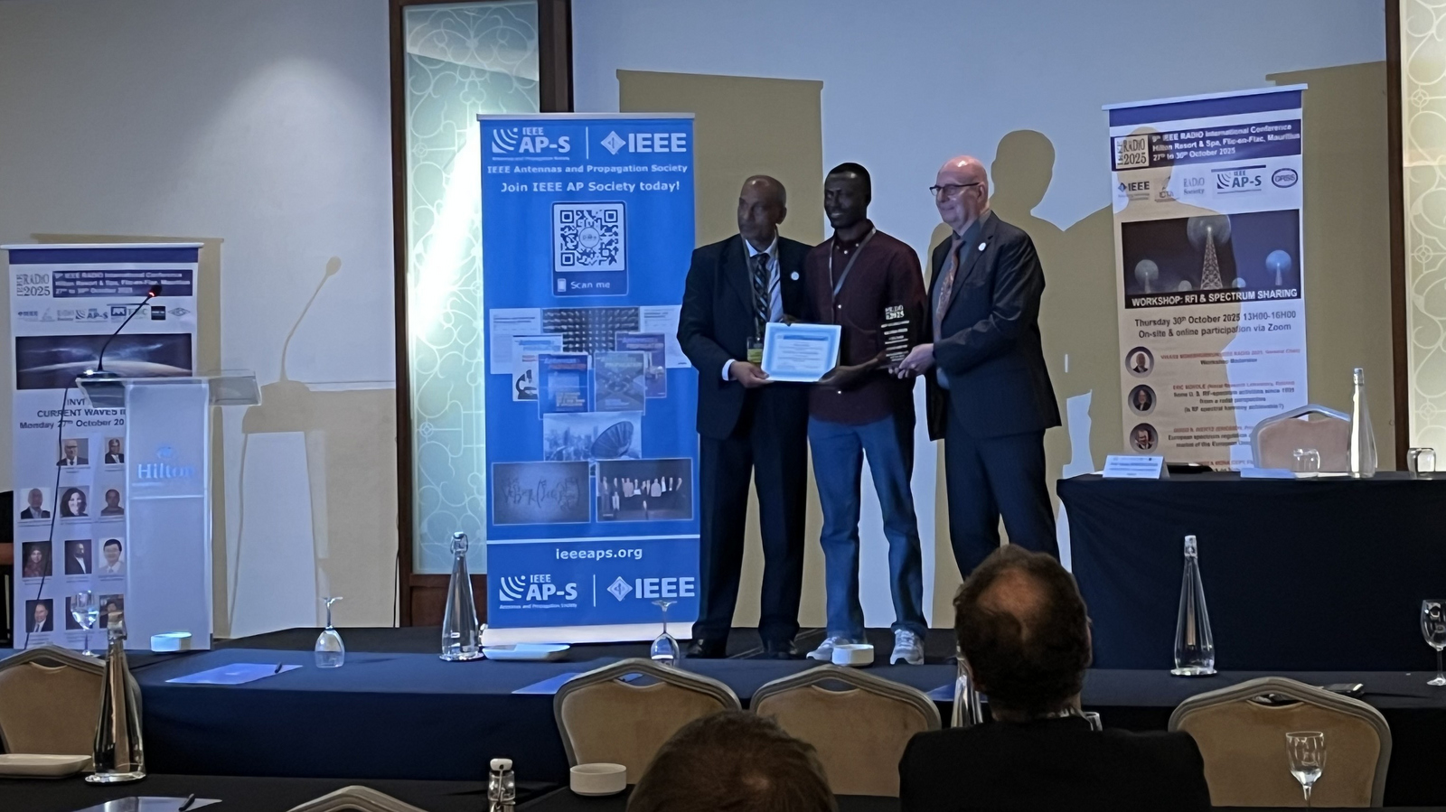 Drei Männer stehe zwischen Roll-ups auf einer Bühne der Mittlere erhält eine Urkunde