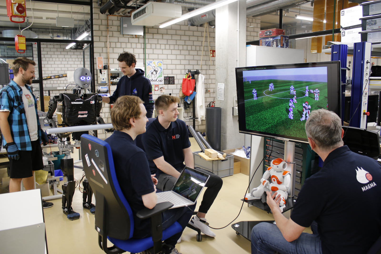 Team Magma sitzt vor einem Bildschirm auf dem ein feld mit simulierten Robotern zu sehen ist, im Hintergrund steht Sweaty und stemmt beobachtet von zwei Teammitgliedern Gewichte